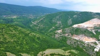 Natural stepped quarry rich in minerals is located near winding intermountain road, against backdrop of Rhodope Mountains and hills with evergreen spruce forests and vegetation. UHD 4K video realtime