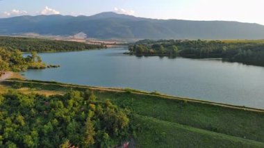 Small blue reflecting lake and clear cool water with stone narrow wild shore in coniferous spruce forest with tall fir trees, against pale blue daytime cloudy sky. UHD 4K video realtime