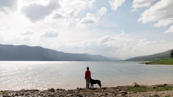 女孩们站在罗特韦勒品种的狗友身边 在清澈的湖畔空旷的野生岩石海岸上 背靠着布满云杉林的罗德尔山脉 — 图库视频影像