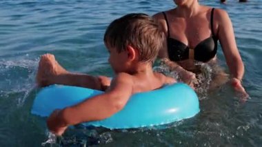 Sarı saçlı mutlu genç anne, küçük neşeli oğluyla oynar ve derin mavi Adriyatik Denizi 'nde parlak mavi şişme bir yüzüğe biner. UHD Video yavaş çekim 4K