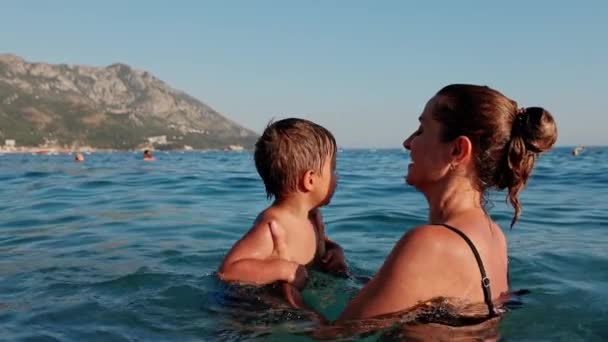 一位穿着游泳衣的快乐的母亲把她的小儿子抱在怀里 在黄昏的阳光下 把他扔到温暖的蓝色亚得里亚海里 盖住她的头 Uhd Video慢动作4K — 图库视频影像