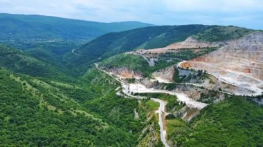 Natural stepped quarry rich in minerals is located near winding intermountain road, against backdrop of Rhodope Mountains and hills with evergreen spruce forests and vegetation. UHD 4K video realtime