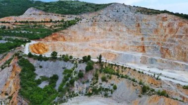 A natural stepped quarry rich in minerals is located near winding road, against backdrop of Rhodope Mountains and hills with evergreen spruce forests and mountain vegetation. UHD 4K video realtime
