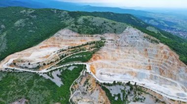 Natural stepped quarry rich in minerals is located near winding intermountain road, against backdrop of Rhodope Mountains and hills with evergreen spruce forests and vegetation. UHD 4K video realtime