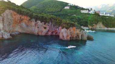 Gezintiler için küçük beyaz bir motorlu tekne yüksek tehlikeli uçurum yamaçlarının altında turkuaz deniz suyunda yüzüyor. UHD Video gerçek zamanlı 4K