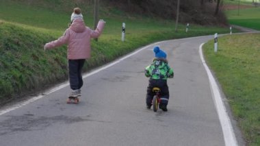 Kaykaylı bir kız kardeş ve erkek kardeşi asfalt yolda bisiklet sürerken kırsal alanda, soğuk bahar havasında yeşil çayırlar arasında. 4K UHD video gerçek zamanlı