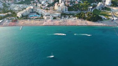 Budva ve Becici tatil beldeleri, sakin Adriyatik Denizi yakınlarında lüks oteller ve rahat plajlarla Karadağ Dağları ve mavi gökyüzüne karşı. UHD Video gerçek zamanlı 4K