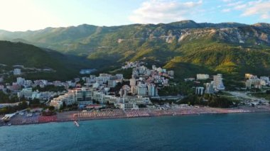 Geniş plaj, güneş şemsiyeleri, plastik beyaz güneşlikler ve Adriyatik Denizi yakınlarındaki Becici tatil beldesinde dinlenen insanlar akşam güneşli gökyüzüne karşı. UHD Video gerçek zamanlı 4K