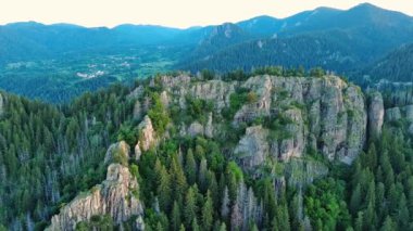 High rocky wild mountain range of Rhodope Mountains covered with green vegetation, against backdrop of mountain valley covered with dense dark spruce forests and blue cloudy sky. UHD 4K video realtime