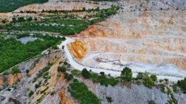 A natural stepped quarry rich in minerals is located near winding road, against backdrop of Rhodope Mountains and hills with evergreen spruce forests and mountain vegetation. UHD 4K video realtime