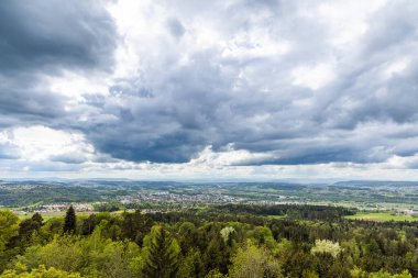 Yoğun bir yeşil orman manzarası, tarım tarlaları ekilmiş ve bulutlu gri gökyüzüne karşı vahşi alçak tepelerde köy evleri.