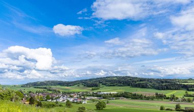 Yoğun bir yeşil orman manzarası, tarım tarlaları ekilmiş ve bulutlu gri gökyüzüne karşı vahşi alçak tepelerde köy evleri.