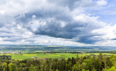 Yoğun bir yeşil orman manzarası, tarım tarlaları ekilmiş ve bulutlu gri gökyüzüne karşı vahşi alçak tepelerde köy evleri.