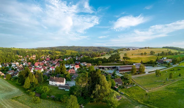 Panorama of a small farming European village with winding streets and cozy residential buildings, green spring tall trees against the background of a dense mixed forest and a cloudy sky