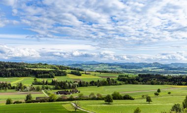 Yoğun bir yeşil orman manzarası, tarım tarlaları ekilmiş ve bulutlu gri gökyüzüne karşı vahşi alçak tepelerde köy evleri.