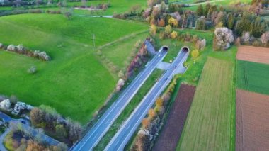 Otoyolları ve yolları olan küçük kırsal Avrupa kasabası, tarım arazileri, ormanlı karanlık tepe siluetleri ve güneşli bir akşam gökyüzü karşısında çiftçi otlakları. Video 4k UHD eş zamanlı