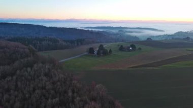 Geniş ve dolambaçlı otoyollar, koyu yoğun çam ormanları, çimenli yeşil çayırlar ve güneşli akşam ışığında beyaz sisle kaplanmış küçük bir köy ile yuvarlanan bir vadiden geçer. Video 4k UHD eş zamanlı