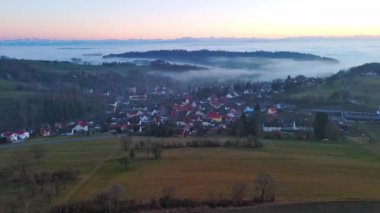 Geniş ve dolambaçlı otoyollar, koyu yoğun çam ormanları, çimenli yeşil çayırlar ve güneşli akşam ışığında beyaz sisle kaplanmış küçük bir köy ile yuvarlanan bir vadiden geçer. Video 4k UHD eş zamanlı