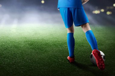 Mavi formalı bir futbolcunun futbol sahasında topa vuruşunun arka görüntüsü.
