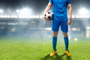 Mavi formalı bir futbolcu futbol sahasında duruyor ve topu tutuyor.
