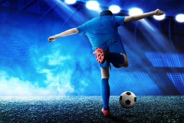 Mavi formalı bir futbolcunun futbol sahasında topa vuruşunun arka görüntüsü.
