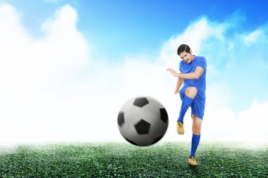 Asyalı futbolcu mavi formalı adam futbol sahasında topa vuruyor.