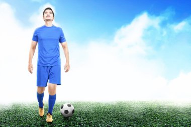 Asyalı futbolcu mavi formalı adam elinde topla futbol sahasında duruyor.