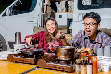 Asyalı çift barbekü yaparken oturmuş doğada kamp faaliyetlerinin keyfini çıkarıyorlar.