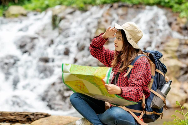 戴着帽子和背包的亚洲女人拿着地图 带着瀑布背景环视四周 — 图库照片