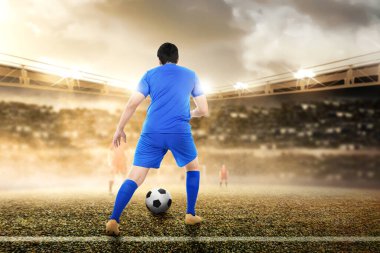 Mavi formalı Asyalı futbolcunun futbol sahasında top sürüşü.
