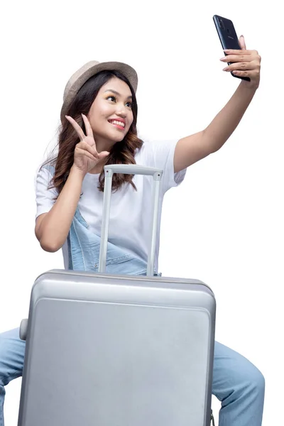 亚洲女人头戴一顶帽子坐在行李箱上 拿着自己的肖像 用白色背景隔离的手机 — 图库照片