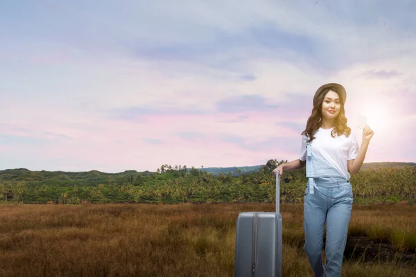帽子をかぶったアジアの女性と日没の空の背景と草原のフィールドに紙を保持しながらスーツケースを運ぶ ストック画像