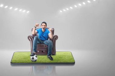 Asyalı adam koltukta oturmuş heyecanlı bir ifadeyle cep telefonu ekranında futbol seyrediyor.