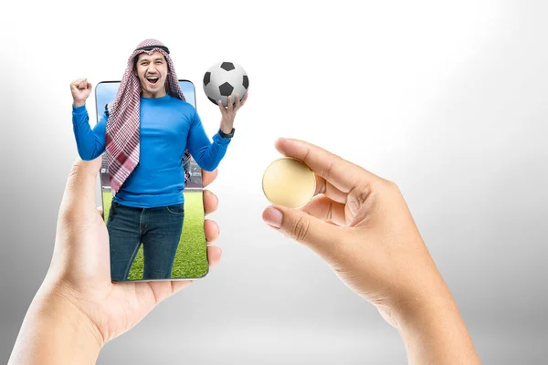 スクリーン上のサッカースタジアムで興奮した表情でボールを保持しながらKeffiyeh立ってアジアの男のビューでコインや携帯電話を保持人間の手 — ストック写真