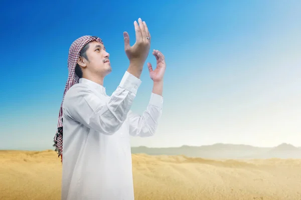 Muslim man with keffiyeh and agal raised hands and praying with blue sky background