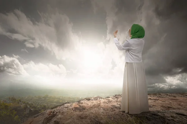 The rear view of a Muslim woman in a veil raising a hand and praying in the dramatic sky background