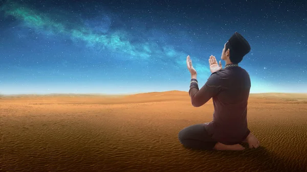 Muslim man sitting while raised hands and praying in the desert