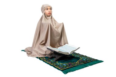 Asian Muslim girl in a hijab with Quran on prayer rug raised hands and praying isolated over white background
