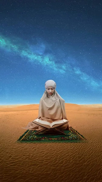 Asian Muslim girl in a hijab with Quran on the prayer rug raised hands and praying in the desert