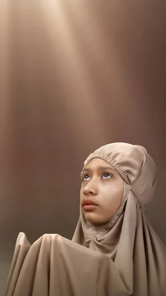 Asian Muslim girl in a hijab raising her hands and praying with light background
