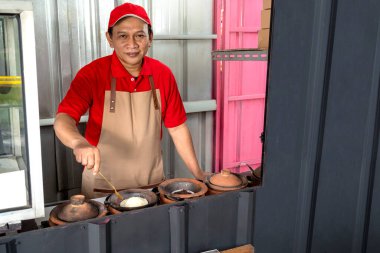 Asian man with a hat and apron cooking serabi in the shop. Serabi is a traditional pancake from Indonesia