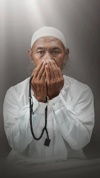 穆斯林男子 留着胡子 头戴白帽 手上拿着祈祷珠子 坐在戏剧性的背景下祈祷 — 图库照片