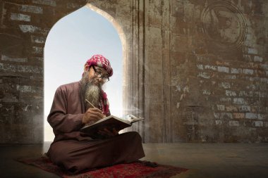 Sakallı Müslüman bir adam camideki kilimin üzerinde oturmuş Kuran okuyor ve ağdalı bir şekilde keffiyeh giyiyor.
