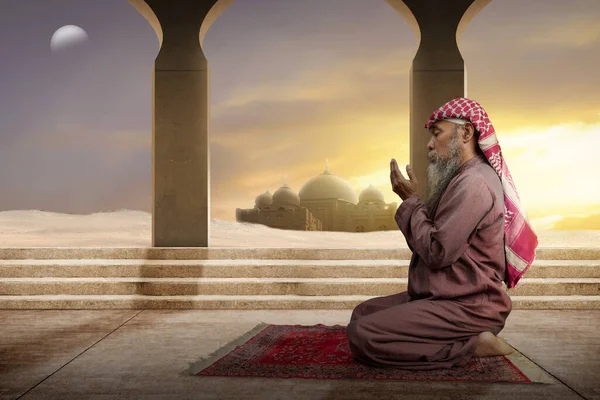 髭を生やしたイスラム教徒の男はモスクの背景と手を上げながら祈りの中でアガルとKefiyehを身に着けています — ストック写真