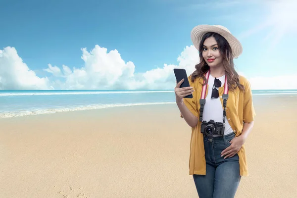 帽子とカメラを持つアジアの女性青い空を背景にビーチを旅する携帯電話を保持 — ストック写真