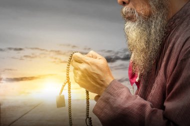 Sakallı, sakallı, sakallı, elinde dua boncuklarıyla dua eden, günbatımı sahnesi arka planında olan Müslüman bir adam.