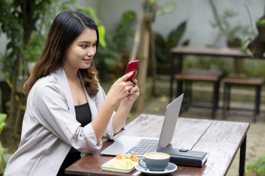 Asyalı iş kadını, kafedeki masada dizüstü bilgisayarı, defteri ve kahve fincanı olan bir cep telefonu kullanıyor.