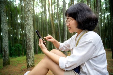 Asyalı bir kadın ormanda cep telefonu kullanarak bankta oturuyor. Seyahat etkinliği
