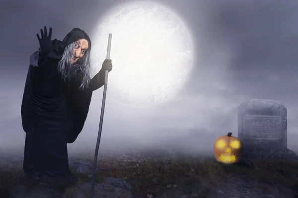 穿着斗篷 拿着棍子的老巫婆站在墓地 背景是夜景 可怕的女巫万圣节概念 — 图库照片