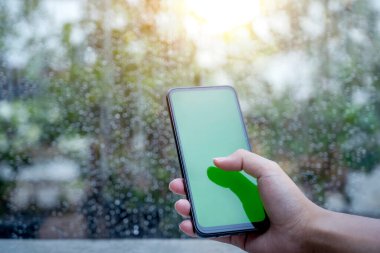 İnsan eli, temiz bir penceresi ve yağmur damlası arkaplanı olan boş ekranlı bir cep telefonu kullanıyor. Kopyalama alanı için boş ekran